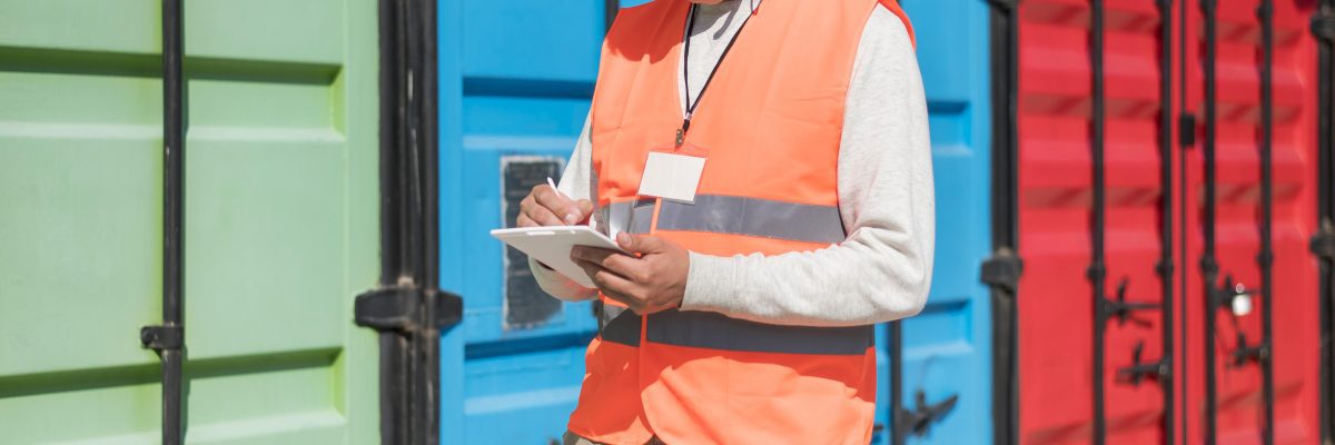 A person wearing a high-visibility orange vest with reflective stripes is holding a clipboard and writing on it with a pen. They are standing in front of large, colorful shipping containers, one green one blue, and the other one is red. The individual’s face is pixelated for privacy. This scene appears to be related to logistics or shipping industry operations, highlighting inventory management or inspection tasks.